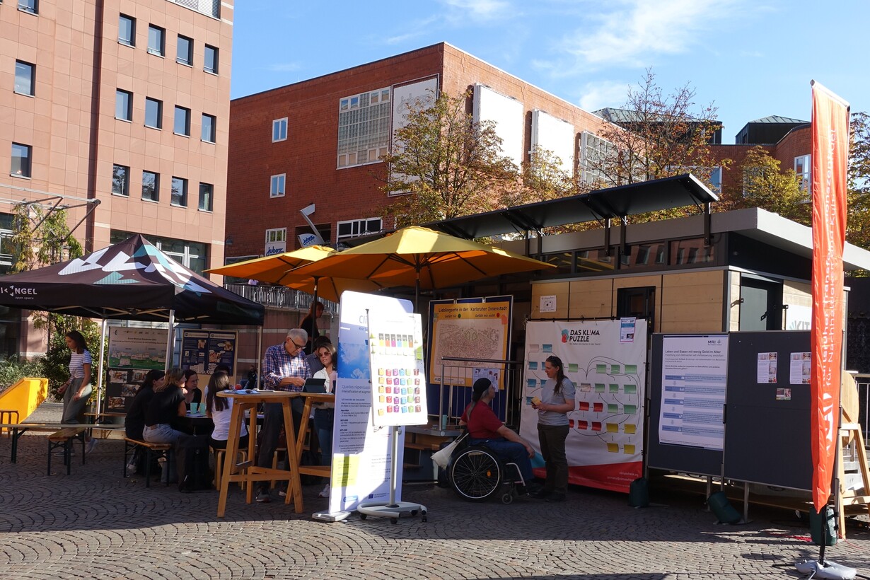 Informationsstand im Freien mit Plakaten und Gesprächen bei sonnigem Wetter in der Stadt.