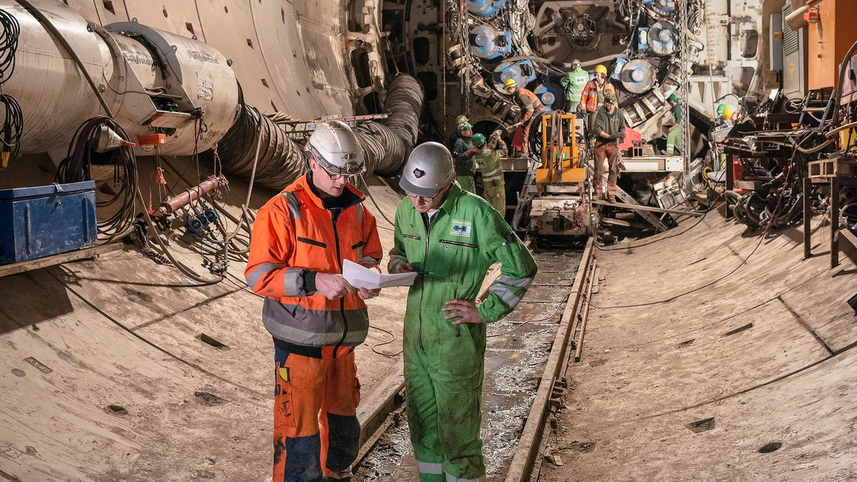 Arbeiter in Schutzkleidung besprechen Pläne im Tunnelbau mit Tunnelbohrmaschine im Hintergrund.