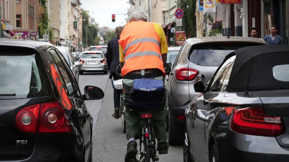 Radfahrer in Warnweste fährt zwischen parkenden Autos auf einer Stadtstraße.