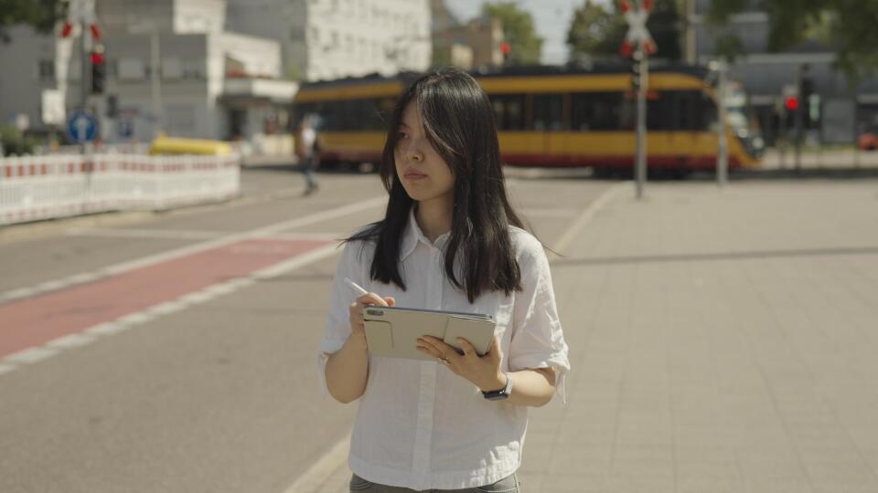 Frau steht mit Tablet und Stift auf Bürgersteig vor Straßenbahn und Ampel bei Sonnenschein.